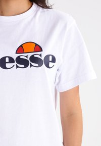 Hvit bomull t-skjorte med ordet "esse" i fet svart skrift, med en stilisert grafikk i rødt og oransje over teksten. Korte ermer.