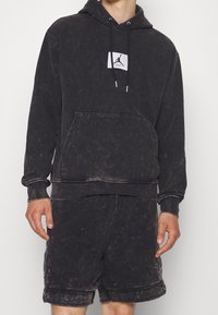 Felpa con cappuccio nera in cotone, dotata di tasca frontale, cappuccio con stringa e una toppa logo bianca; abbinata a pantaloni corti neri con un design simile.