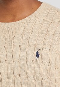 Beżowy sweter z dzianiny w splot warkoczowy, z prążkowanym okrągłym dekoltem i małym granatowym logo na lewej piersi. Teksturalny, miękki materiał.