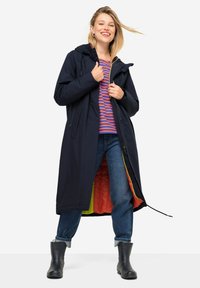 Cappotto lungo impermeabile blu navy con cappuccio, dotato di un luminoso rivestimento interno arancione, due tasche frontali e orlo con cordino. Camicia a righe sotto.