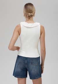 Top bianco senza maniche con colletto arricciato, texture a coste. Abbinato a pantaloni shorts in denim scuro con tasche posteriori e taglio dritto.