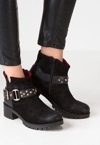 Black ankle boots made of suede with a leather strap featuring gold studs, a side zipper, and a textured rubber sole.