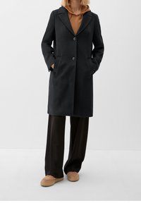 Svart ullblandad kappa med slagkrage, två framfickor och två knappar. Buren över en brun huvtröja och matchad med svarta vida byxor.