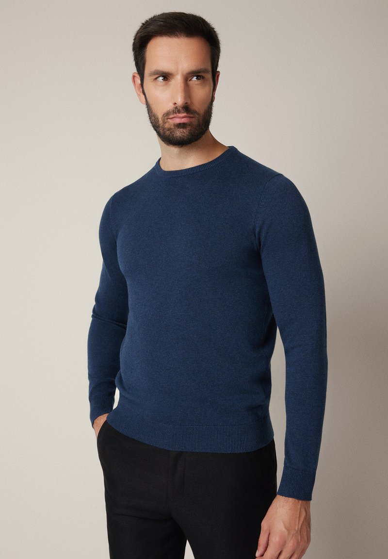 Granatowy długorękawowy sweter o miękkiej fakturze, z klasycznym okrągłym dekoltem i ściągaczami przy mankietach, zestawiony z ciemnymi spodniami.