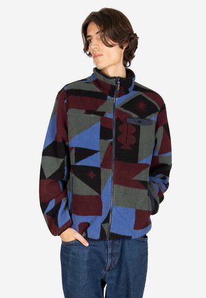 Junger Mann, der eine mehrfarbige Fleecejacke mit geometrischem Muster und blaue Jeans trägt, schaut seitlich mit den Händen in den Taschen.