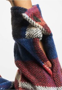 Vlněná flanelová košile s kostkovaným vzorem v červené, modré a béžové barvě. Obsahuje černé zapínání na knoflíky a měkkou, strukturovanou látku.