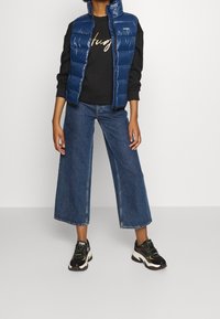 Blauwe bodywarmer over een zwarte sweatshirt met gouden tekst, gecombineerd met wijde blauwe jeans en zwarte sneakers met groene accenten.