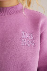 Ljubičasti dukserica s rebrastim okruglim izrezom, mekanom teksturom i izvezenim natpisom "DaNcE" sa zvezdastim akcentima u svetlijoj nijansi ljubičaste.
