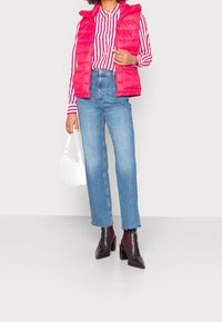 Gilet rembourré rose par-dessus une chemise rayée rouge et blanche, associé à un jean droit bleu clair et des bottines bordeaux, tenant un sac à main blanc.