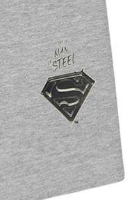 Graues T-Shirt mit einem glänzenden schwarzen Superman-Logo mit diamantartigem Umriss und dem Text "Man of Steel" in stilisierter Schrift darüber.