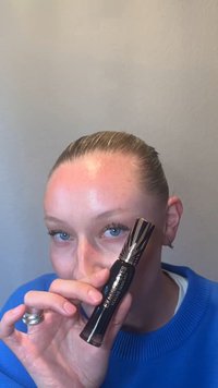 Person with light skin and blue eyes holds a black tube of volume mascara close to their face, wearing a blue sweater.