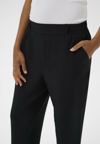 Zwarte broek met een gladde textuur, voorzien van een relaxte tailleband met elastiek en zijzakken, weergegeven met een hand op de zak.