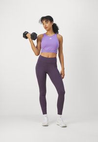Top corto atlético púrpura con el logo de Adidas, combinado con leggings de tiro alto púrpura oscuro que tienen bolsillos laterales, y zapatillas deportivas blancas.