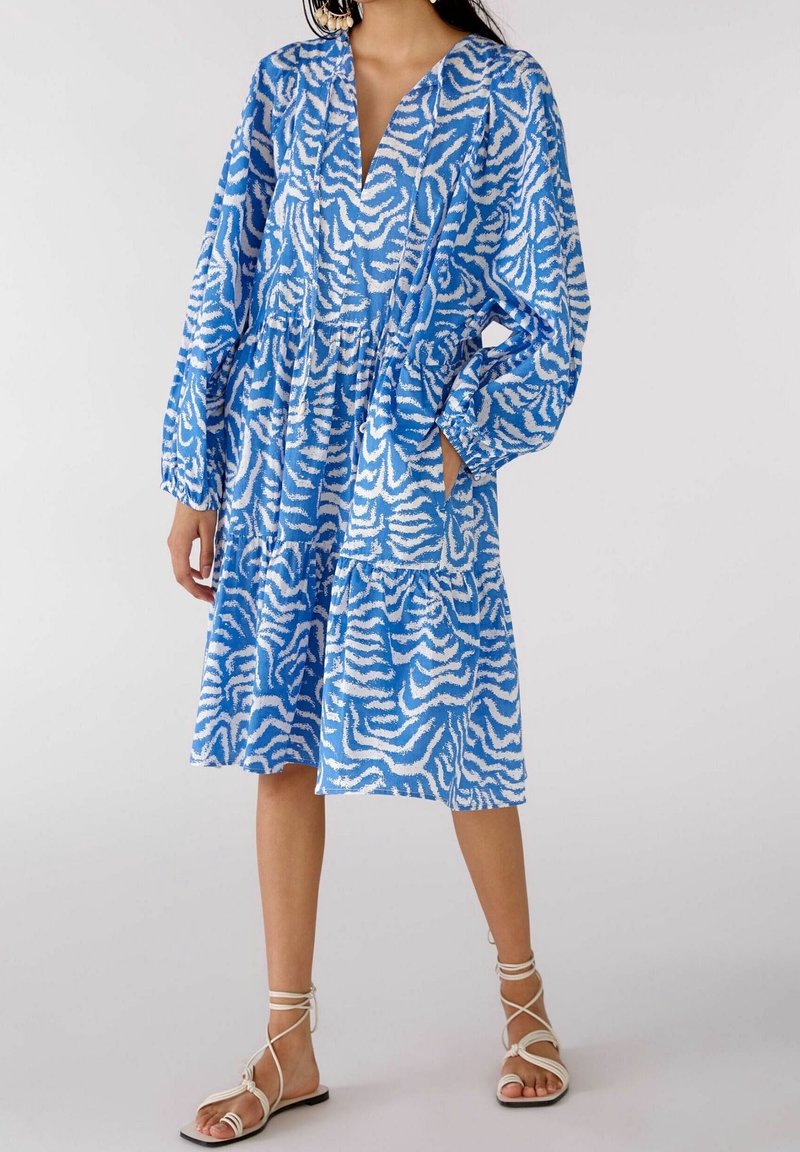 Femme portant une robe bleu et blanc à motifs jusqu’aux genoux avec des manches longues et des sandales plates blanches à brides sur un fond neutre.