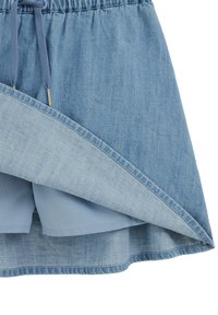 Jeans krilo z elastičnim pasom in vrvico za zavezovanje. Svetlo modri detajl in vidne notranje kratke hlače, ki imajo gladko teksturo in večplastno zasnovo.