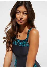 Maillot de bain noir avec un motif pixelisé turquoise au niveau du col. Texture lisse avec de larges bretelles, mettant en valeur une coupe ajustée.