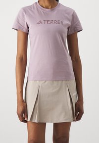 Kvinna som bär en ljuslila TERREX t-shirt och en beige plisserad TERREX kjol, står med avslappnade armar mot en vit bakgrund.