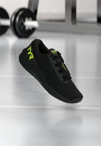 Zapatilla deportiva negra con parte superior de malla, punta redonda y suela texturizada. Presenta marcas y cordones en verde lima. El fondo muestra equipo de gimnasio.