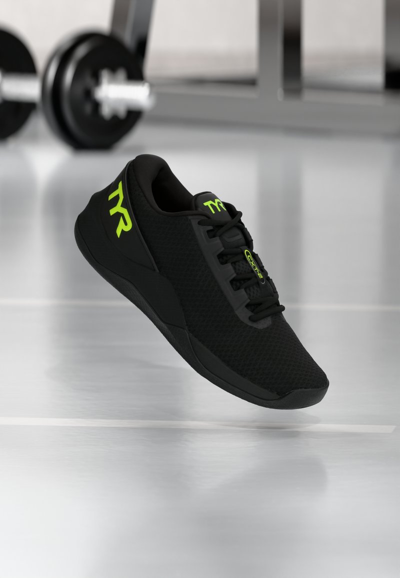 Zapatilla deportiva negra con parte superior de malla, punta redonda y suela texturizada. Presenta marcas y cordones en verde lima. El fondo muestra equipo de gimnasio.