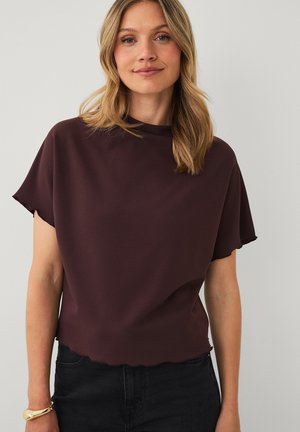 Vrouw met schouderlang blond haar, gekleed in een donkerbruin shirt met korte mouwen en zwarte broek, staand tegen een effen lichte achtergrond.