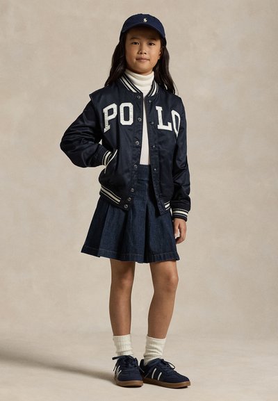 Niña con chaqueta POLO azul marino, gorra a juego, cuello alto blanco, falda plisada azul marino, calcetines blancos y zapatillas azul marino, de pie sobre fondo liso.