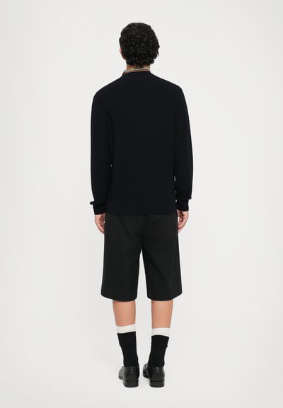 Maglione lungo blu navy con texture e colletto colorato, abbinato a pantaloni corti neri al ginocchio, calze nere e scarpe nere.