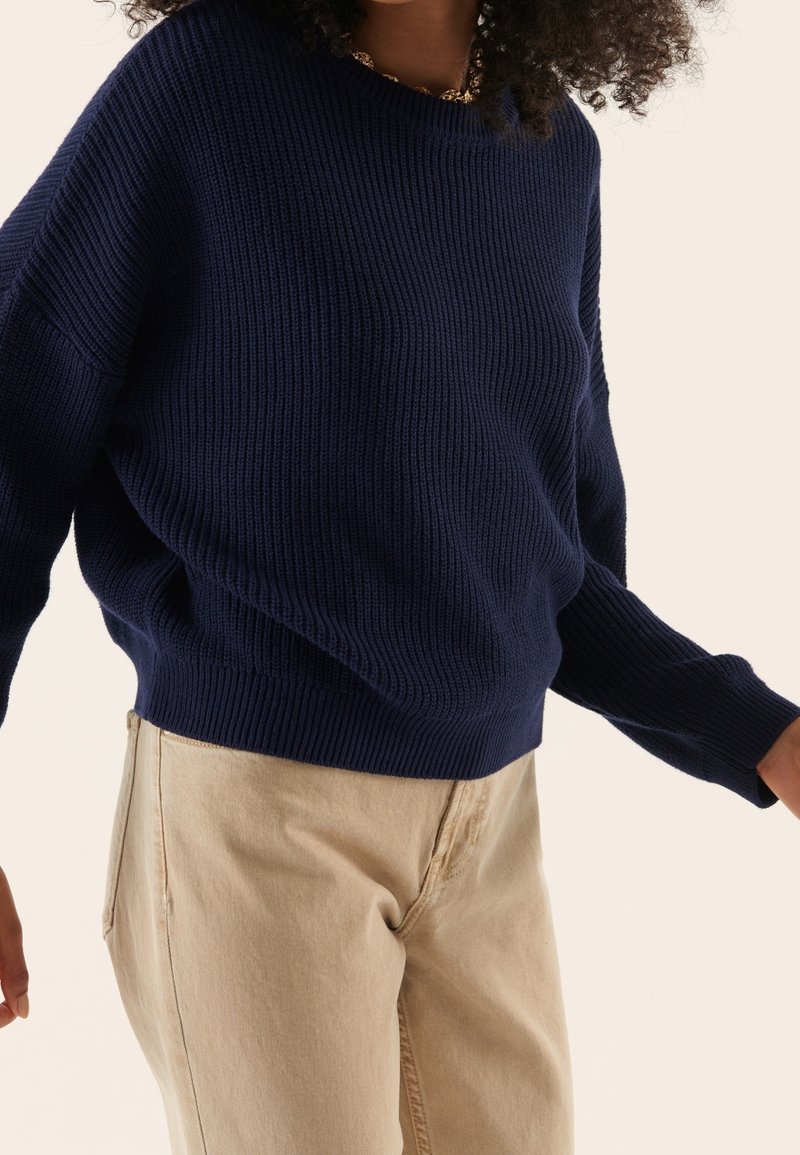 Granatowy sweter o prążkowanej strukturze z szerokim dekoltem typu łódka i obniżonymi ramionami, w zestawieniu z beżowymi spodniami, ukazujący miękką fakturę materiału i luźny krój.