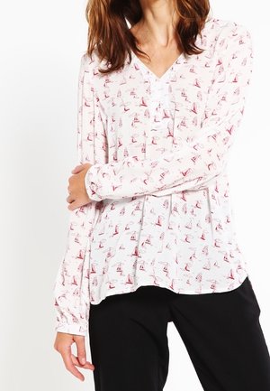 Weiße Bluse mit einem roten Segelboot-Druck, V-Ausschnitt, Knopfdetails und langen Ärmeln, kombiniert mit schwarzen Hosen.