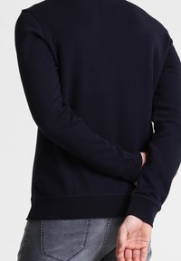 Person in einem schwarzen Langarmshirt mit verschränkten Händen hinter dem Rücken, kombiniert mit grauen Denim-Jeans.