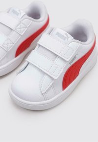 Sneakers bianchi con accenti rossi, dotati di due cinturini in velcro, una tomaia in sintetico liscio e una suola testurizzata. Logo Puma di lato.