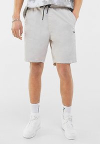 Shorts gris clair avec cordon de serrage, poches latérales, texture douce et logo sur la jambe droite, associés à des sneakers blanches montantes.