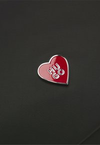 Rød hjerteformet nål med sølvkanter og "HUGO" i sølvbogstaver på tekstureret sort stofbaggrund.