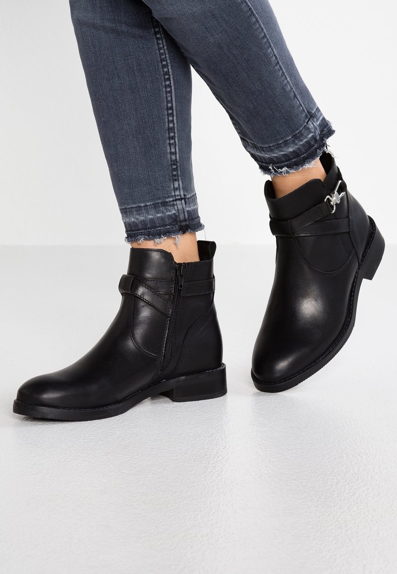 Black leather ankle boots with side zipper, flat heel, and decorative strap. Worn with frayed hem dark jeans, showing foot positioning.