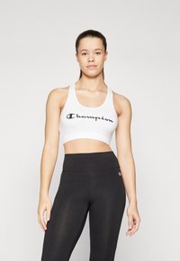 Λευκό αθλητικό σουτιέν με μαύρο λογότυπο "Champion", συνδυασμένο με ψηλόμεσα μαύρα κολάν. Το ύφασμα φαίνεται ελαστικό με ομαλή υφή.