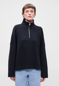 Pulôver preto com fecho em zíper e gola alta, feito de um tecido macio. Apresenta mangas compridas e um corte solto, combinado com jeans azul claro.