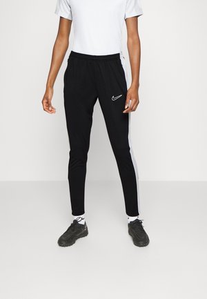 Persoon draagt zwarte Nike trainingsbroek met witte zijstrepen, zwarte sneakers, witte sokken en een wit kort mouw shirt tegen een effen achtergrond.
