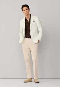 Muž v bielom saku, tmavohnedom tričku, béžových nohaviciach a svetlobežových espadrilkách stojí opretý o jednoliatu sivú stenu.