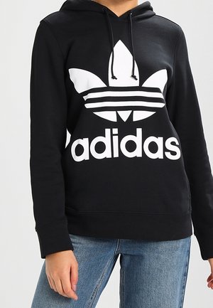 Osoba w czarnej bluzie z kapturem Adidas z dużym białym logo i napisem, w zestawieniu z jasnoniebieskimi dżinsami, stoi na jednolitym tle.