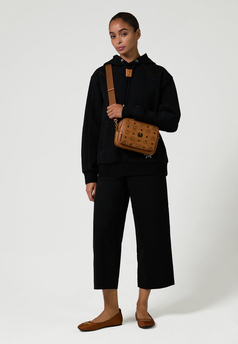 Frau in schwarzem Kapuzenpullover und weit geschnittenen, knöchellangen Hosen, die eine braune Umhängetasche hält und passende braune Flats trägt, steht vor einem schlichten Hintergrund.