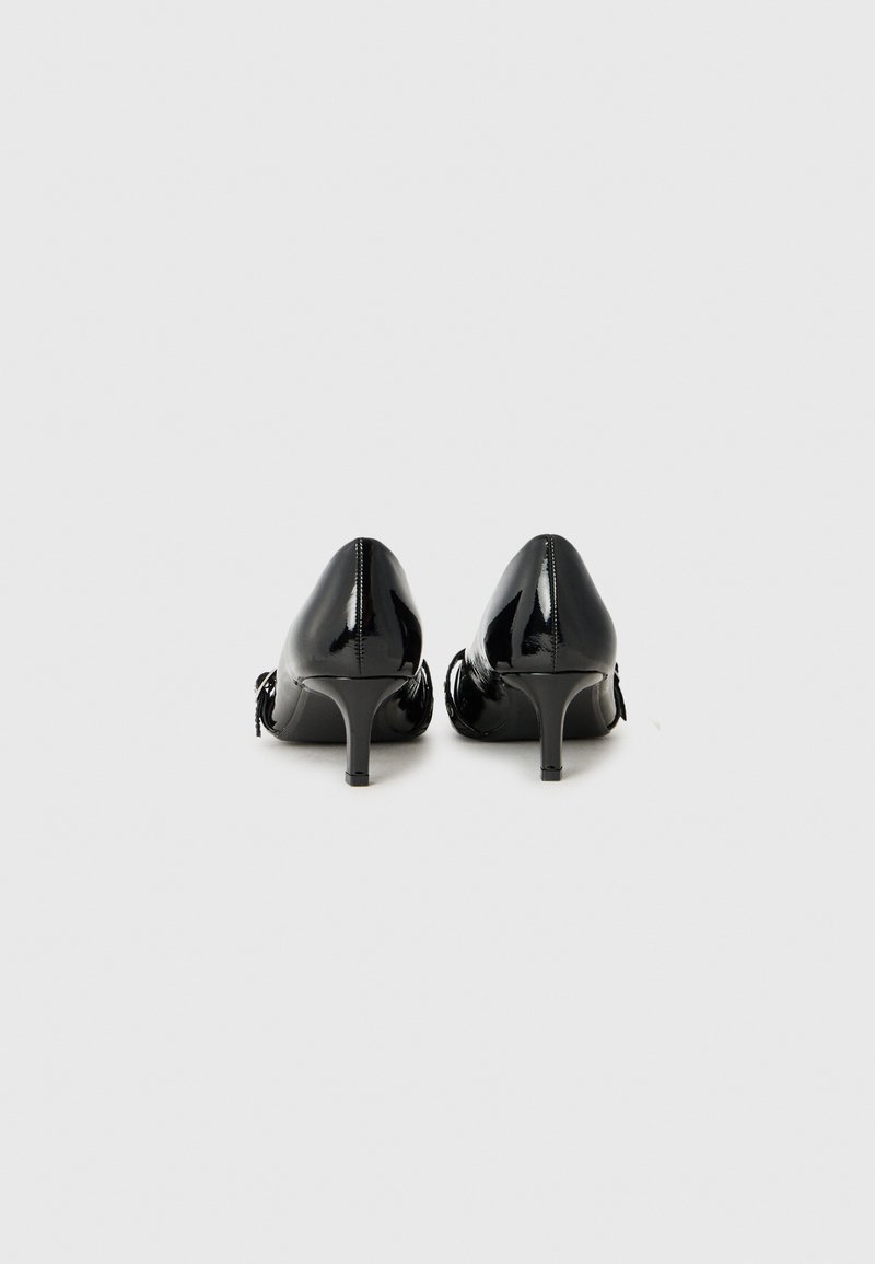 Tacones de charol negros con punta afilada y tacón delgado. Presentan un sutil detalle de volantes en la parte posterior. Acabado suave y brillante.