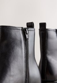 Zwarte leren enkellaarzen met ritssluiting, gladde textuur en contrasterende stiksels. Voorzien van trekbandjes aan de achterkant voor gemakkelijk aan- en uittrekken.