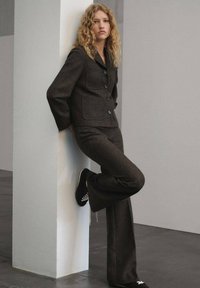 Bruine wollen blazer met grote knopen en zakken, gecombineerd met bijpassende flared broeken; gedragen met zwarte schoenen met witte veters.