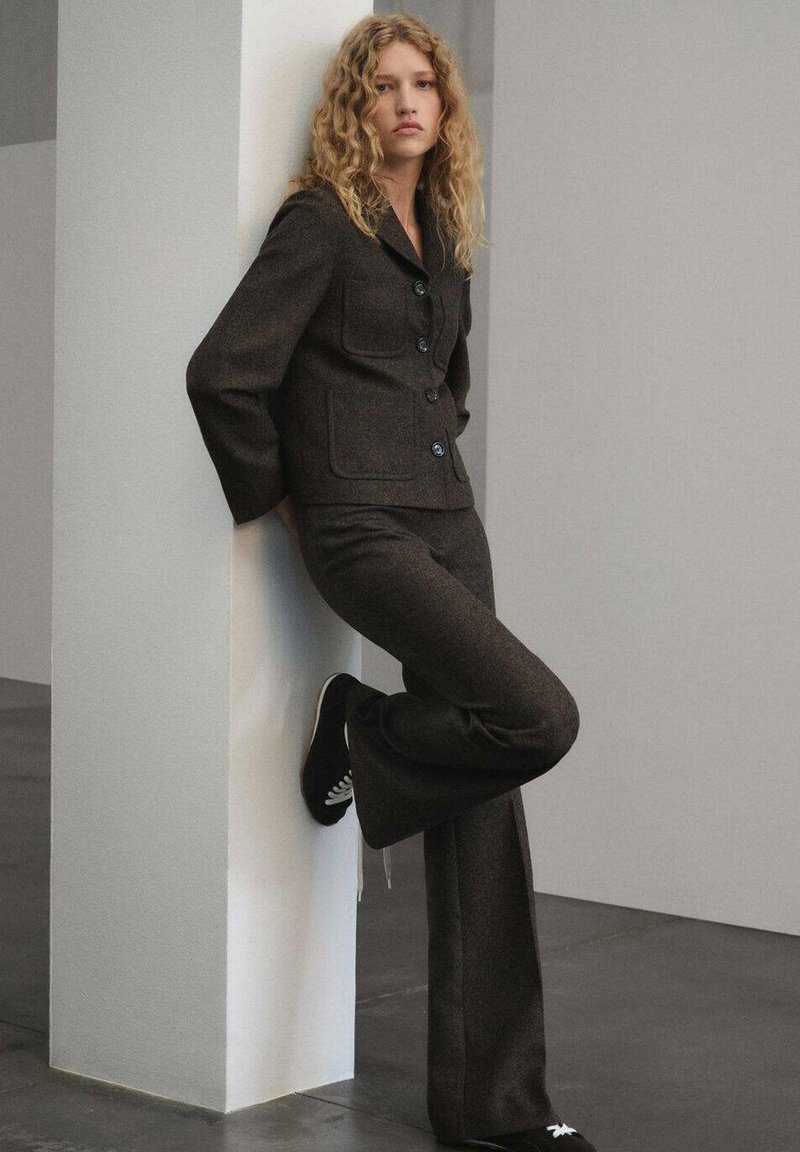 Blazer in lana marrone con grandi bottoni e tasche, abbinato a pantaloni svasati coordinati; indossato con scarpe nere con lacci bianchi.
