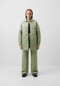 Costum cu quilt, de culoare verde deschis, care include o jachetă cu nasturi și pantaloni lejeri, cu cusături evidente și un mic logo pe jachetă.