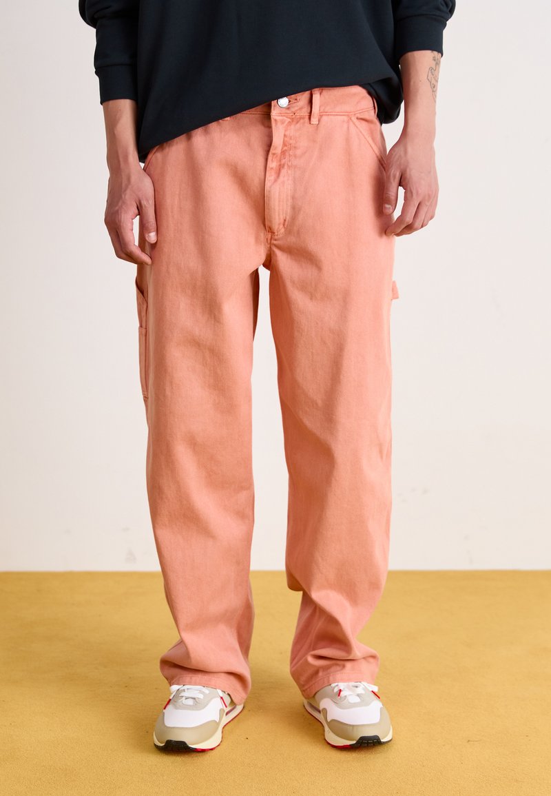 Pink cargo pants made of cotton, featuring side pockets, a relaxed fit, and cuffed hems, paired with light-colored sneakers.