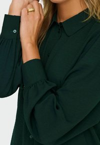 Dunkelgrünes Hemd mit Knopfverschluss aus strukturiertem Stoff, mit Puffärmeln und spitzem Kragen. Enthält ein hellblaues Knopfdetail am Ärmel.