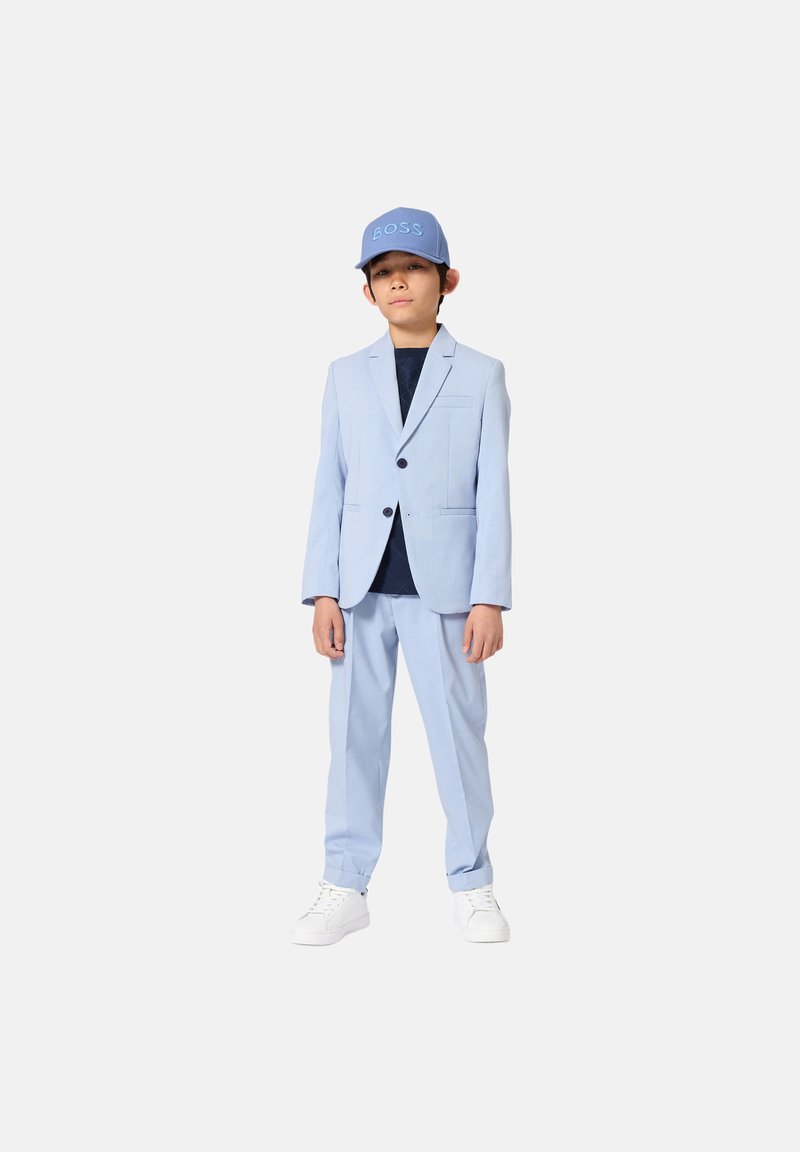 Costume bleu clair avec une veste à un seul bouton et un pantalon assorti. Porté avec une chemise bleu marine et une casquette bleue arborant le logo "BOSS". Baskets blanches.