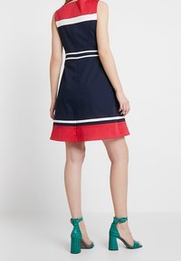 Marinblå klänning med röda och vita ränder; ärmlös design, utsvängt midja, dragkedja i ryggen. Tealfärgade klackar med krokodilmönster och blockklackar.