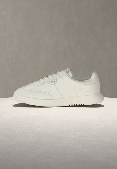 Λευκό χαμηλό sneaker με δετή κορδόνια και διακριτικό λογότυπο στο πλάι, ακουμπισμένο σε λείο φωτεινό υπόστρωμα πάνω σε ουδέτερο φόντο.