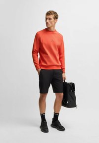 Helles orange Sweatshirt mit Rundhalsausschnitt, schwarze Shorts und schwarze Turnschuhe. Trägt einen schwarzen Rucksack mit aufgerolltem oberen Teil.